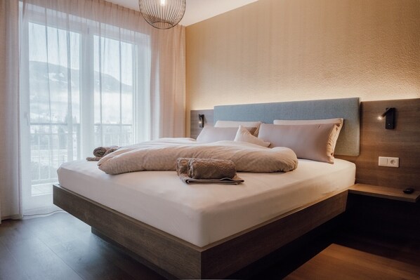 Une chambre d’hôtel moderne avec un grand lit, une table de chevet, une lampe fixée au mur et une vue dégagée à travers des rideaux légers.