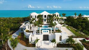 A white, modern resort with a central pool, surrounded by palm trees and a view of the ocean.
