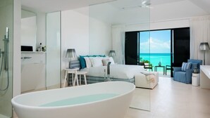 A modern hotel room with a large bathtub, a bed with blue and white bedding, a seating area with a sofa and armchairs, and a view of the ocean through large windows.