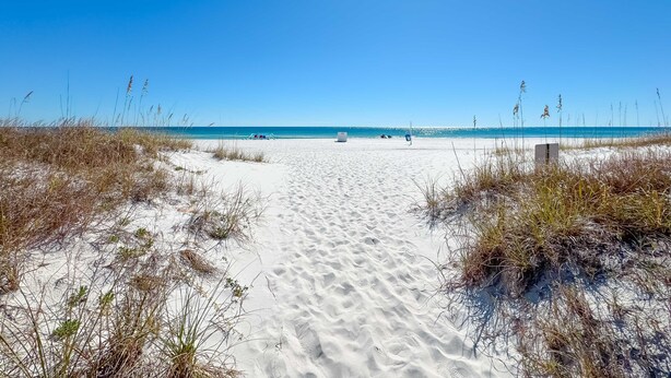 Ein sandiger Küstenpfad führt zum Meer, begleitet von spärlicher Vegetation an beiden Seiten.
