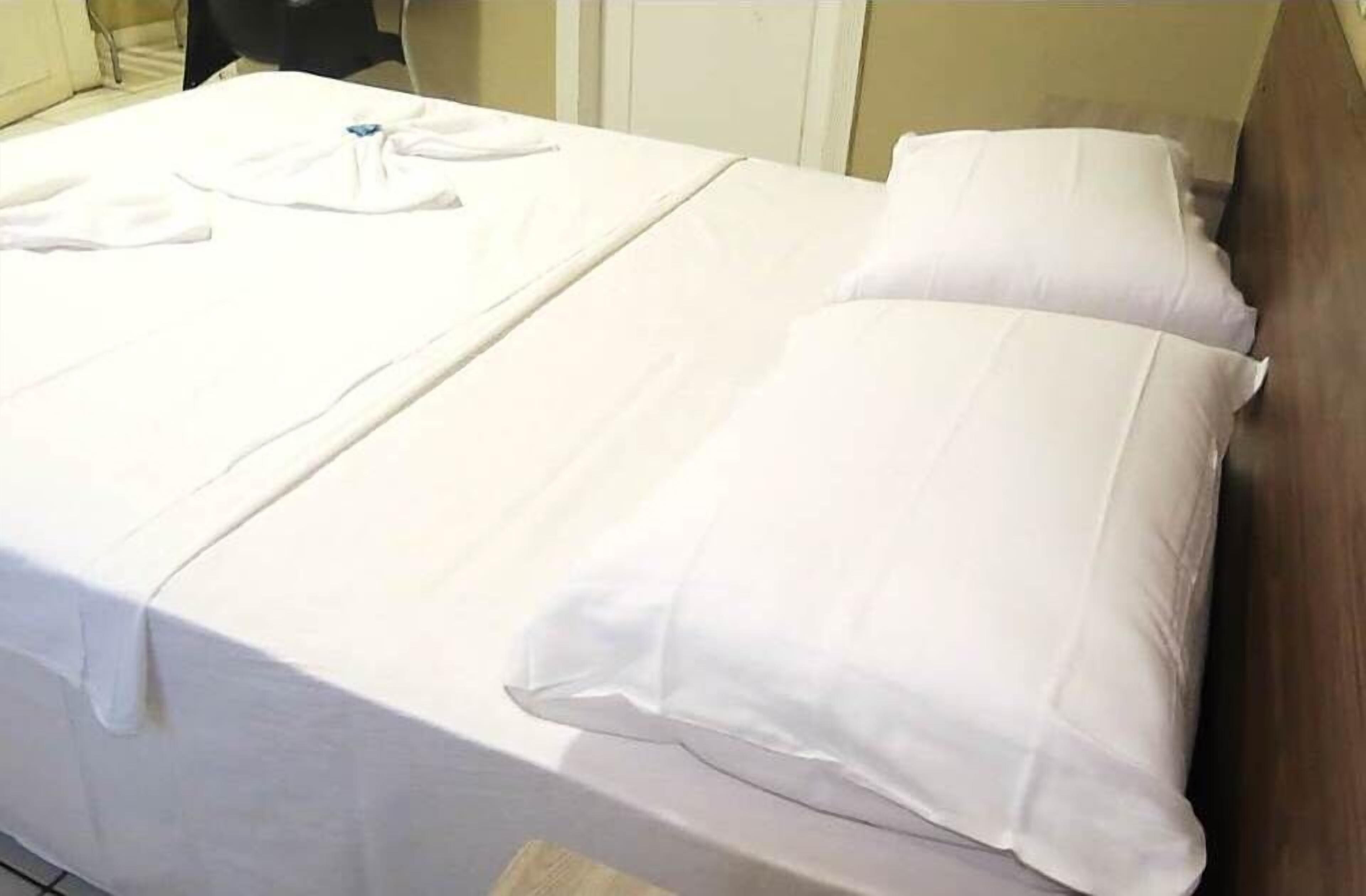 A neatly made bed with white linens and pillows, a folded white towel, and a small table with a white cloth.