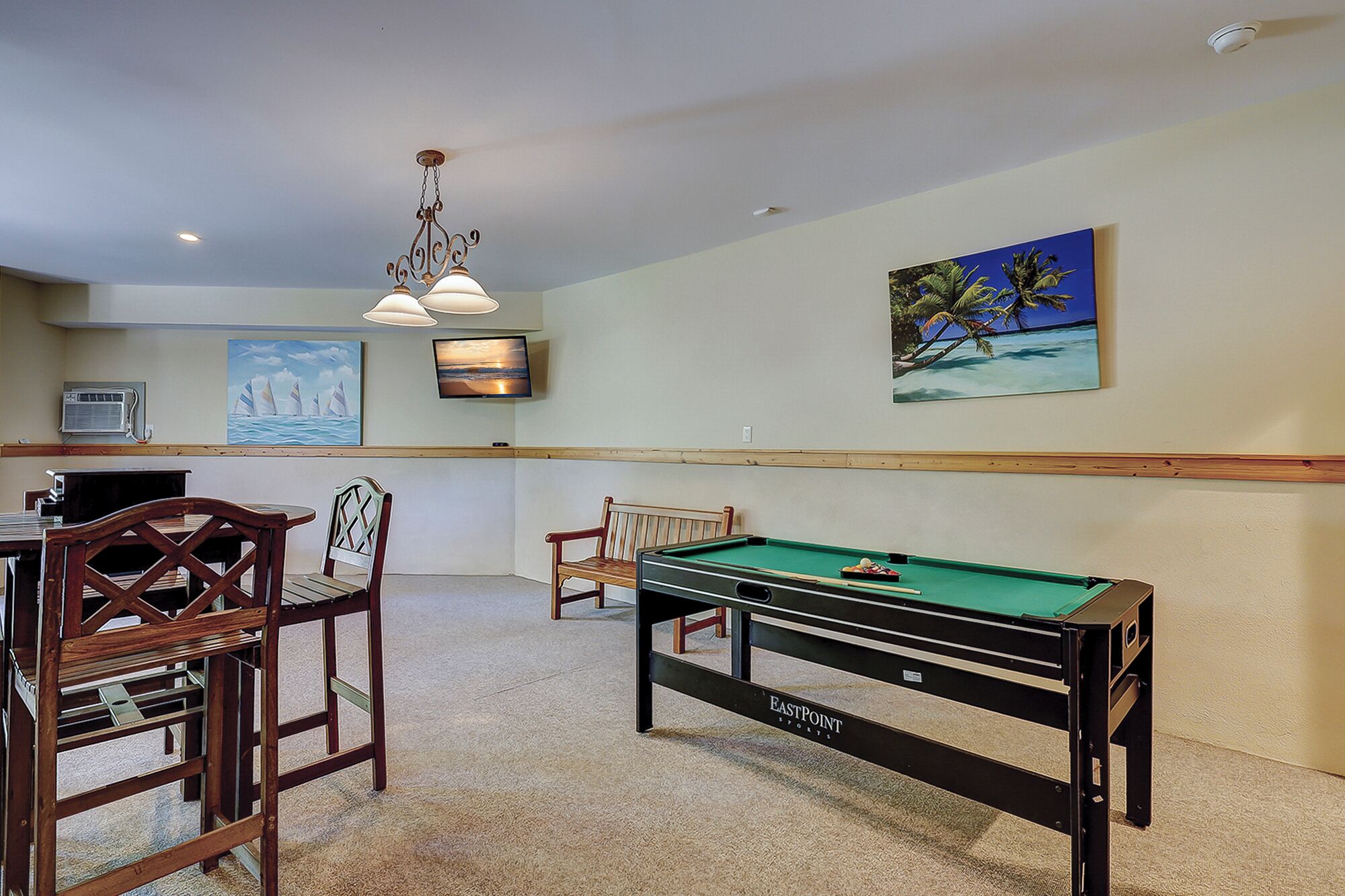 Une table de billard recouverte d’un drap de feutre vert, des chaises en bois, un ventilateur de plafond et des œuvres d’art accrochées au mur, dont une peinture représentant une plage tropicale et un voilier.