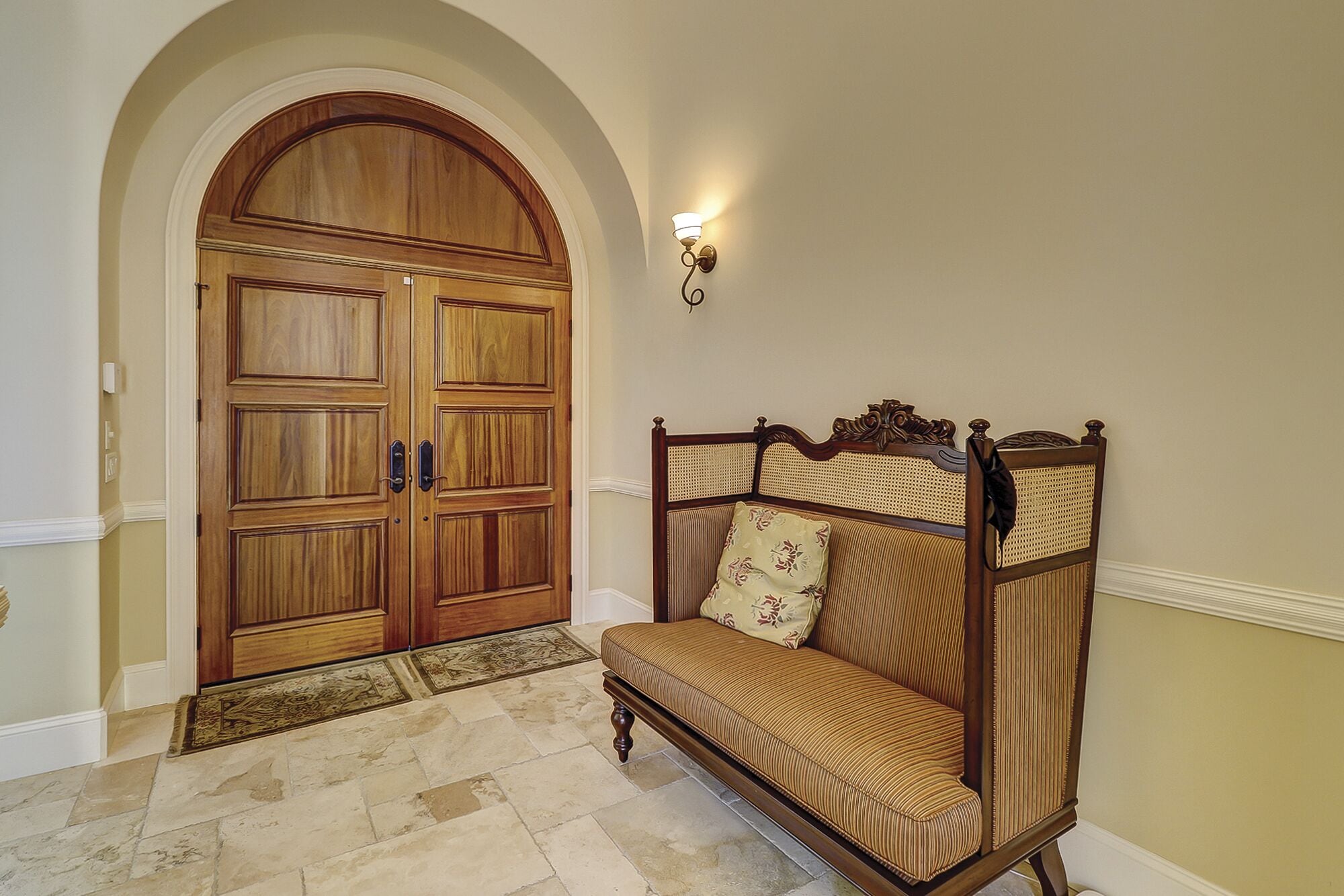 Un pasillo con una puerta doble de madera, un banco con respaldo de mimbre y una lámpara de pared.