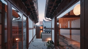 Un couloir de style japonais traditionnel, avec des poutres en bois, un revêtement de sol en tatami et un petit jardin agrémenté de pierres plates servant de pas et d’une lanterne.