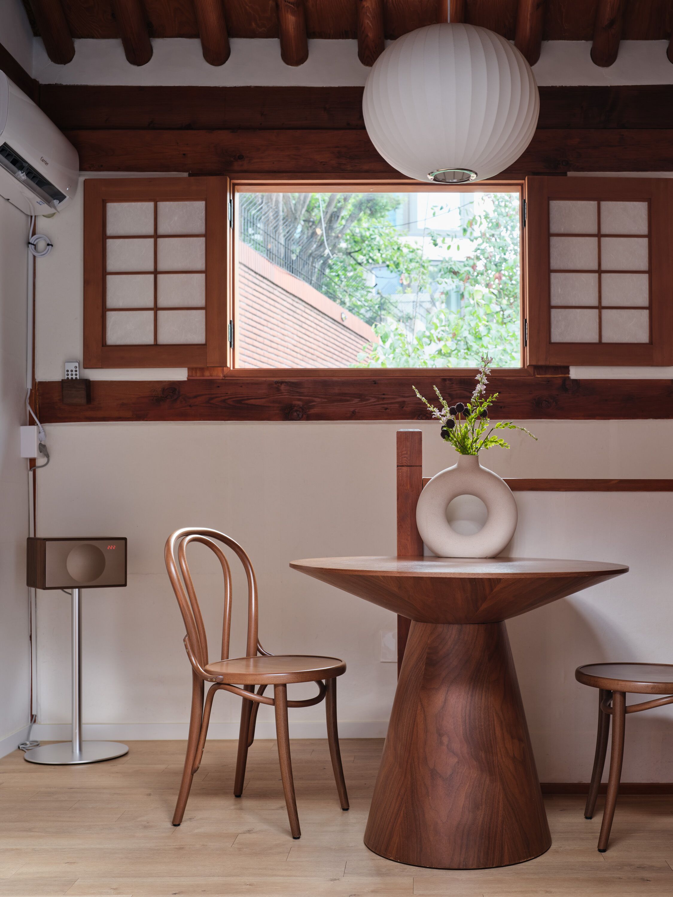 A dining area with a round wooden table, two chairs, a vase with flowers, and a window with a view of greenery.