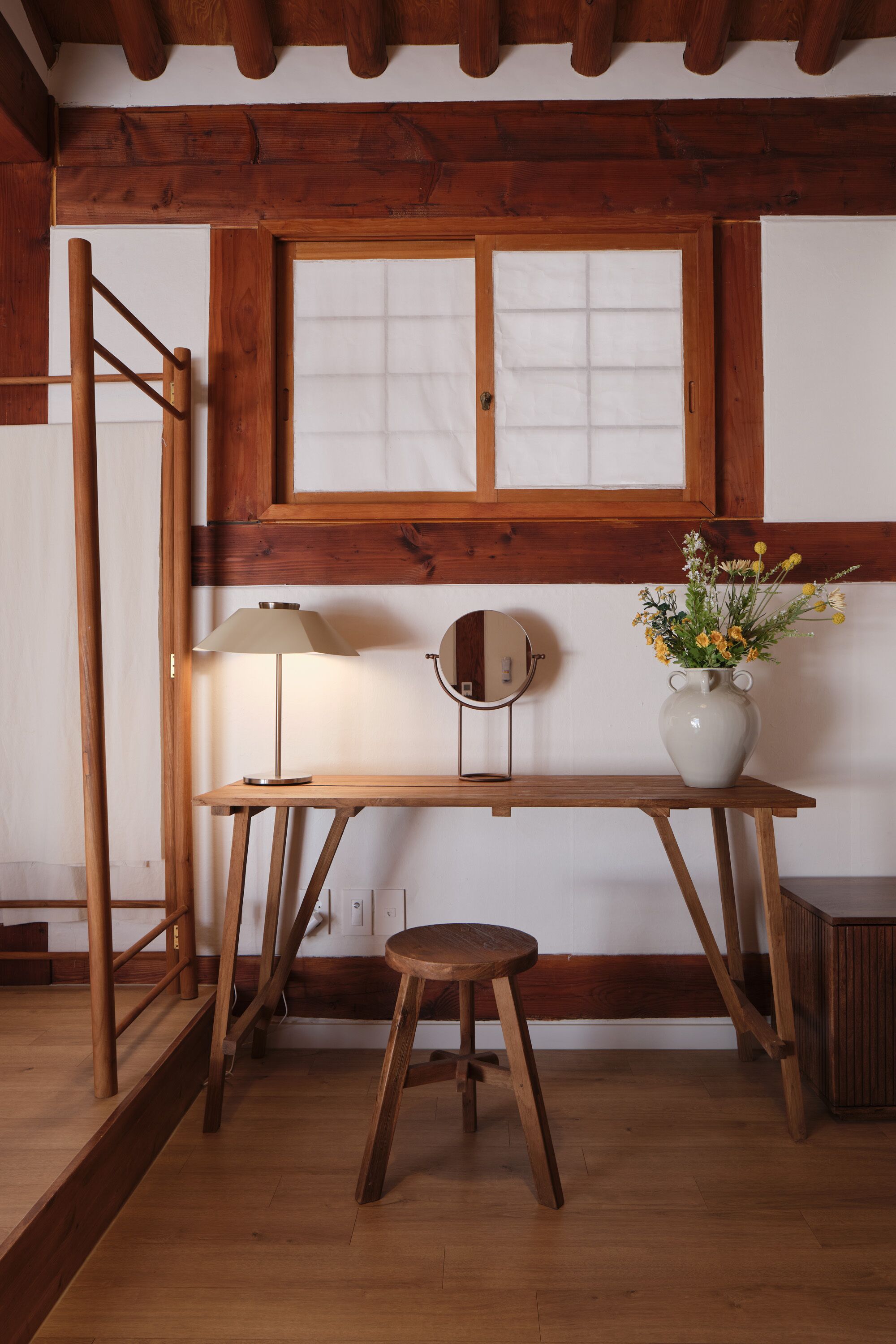 A wooden desk with a lamp, a vase with flowers, and a mirror.
