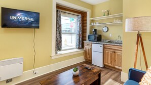 A compact kitchen with a microwave, coffee maker, and a sink. There's a TV mounted on the wall displaying 'Universal 100th Anniversary'. A window with curtains and a view of the street is visible. - Downtown Friday Harbor! Adults Only (CCS#2) (Friday Harbor)