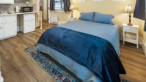 A bedroom with a large bed, two bedside tables with lamps, a window with curtains, a kitchen area with cabinets and appliances, and a rug on the floor.