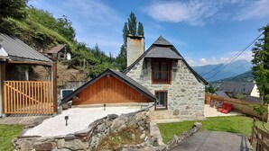 Una casa in pietra con portico in legno e un camino in pietra, circondata da un muro in pietra e un vialetto sterrato.