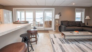 A modern kitchen with a large island, a dining area with a sofa and chairs, and a view of the ocean through the sliding glass doors. - Seneca Memories: Luxury Lakeside Home on Seneca Lake! (Penn Yan)