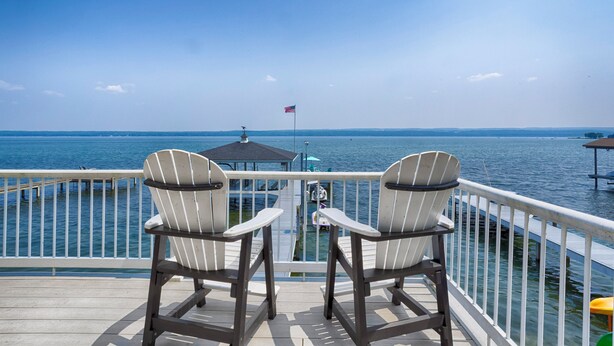 A deck with two Adirondack chairs facing a dock and a pier with a flag. - Seneca Memories: Luxury Lakeside Home on Seneca Lake! (Penn Yan)