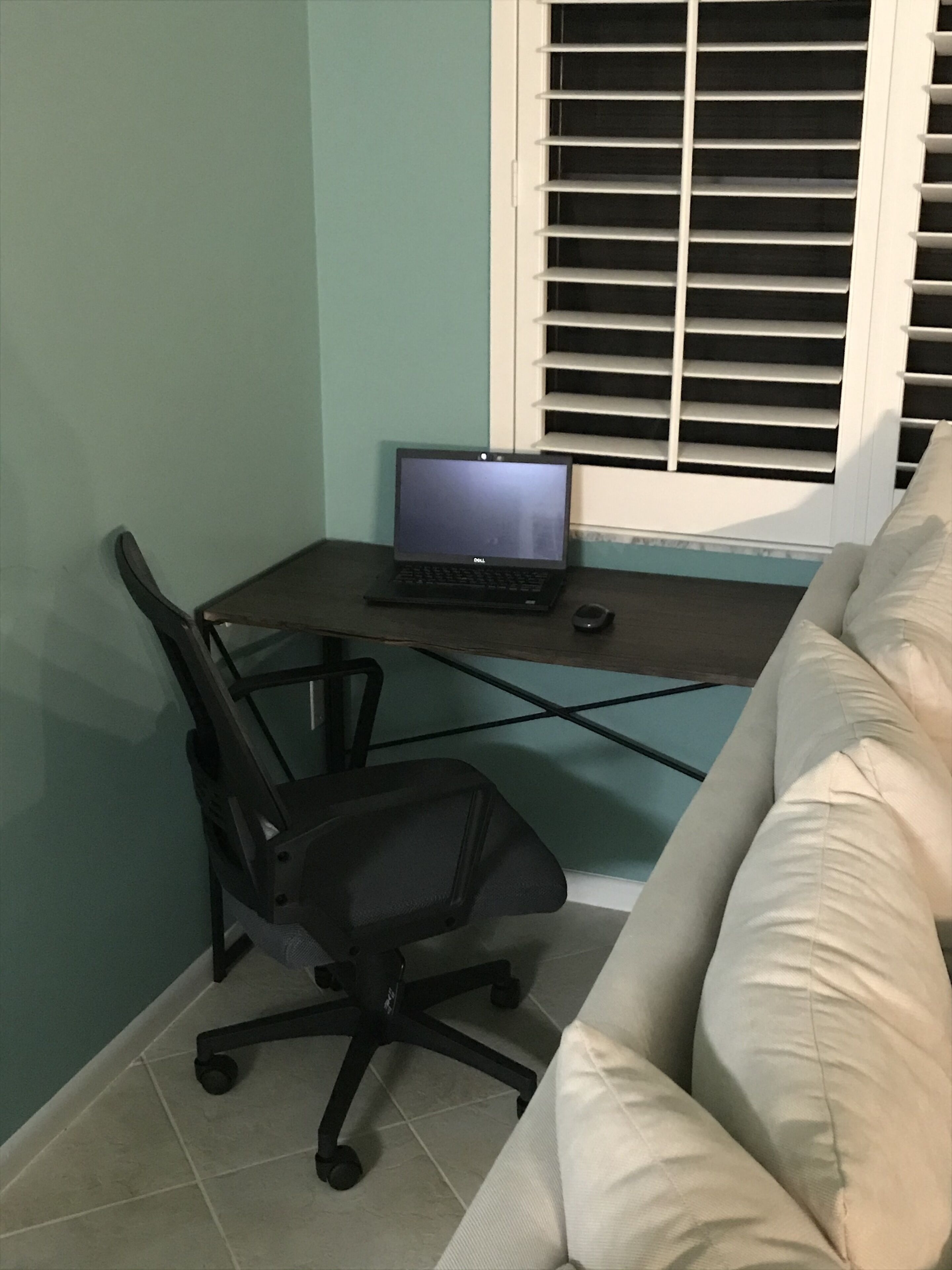 A room with a desk, a laptop, a black office chair, and a window with blinds.
