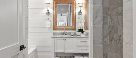 Un baño con paredes de paneles blancos, un espejo con marco de madera, encimeras de mármol y suelo de madera.