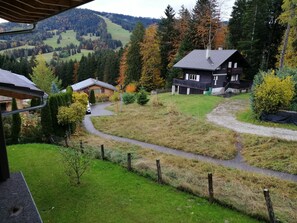 Une vue d’un paysage rural avec deux maisons : l’une a un toit en pente et l’autre, un balcon. Les deux sont entourées d’arbres et d’un sentier sinueux.
