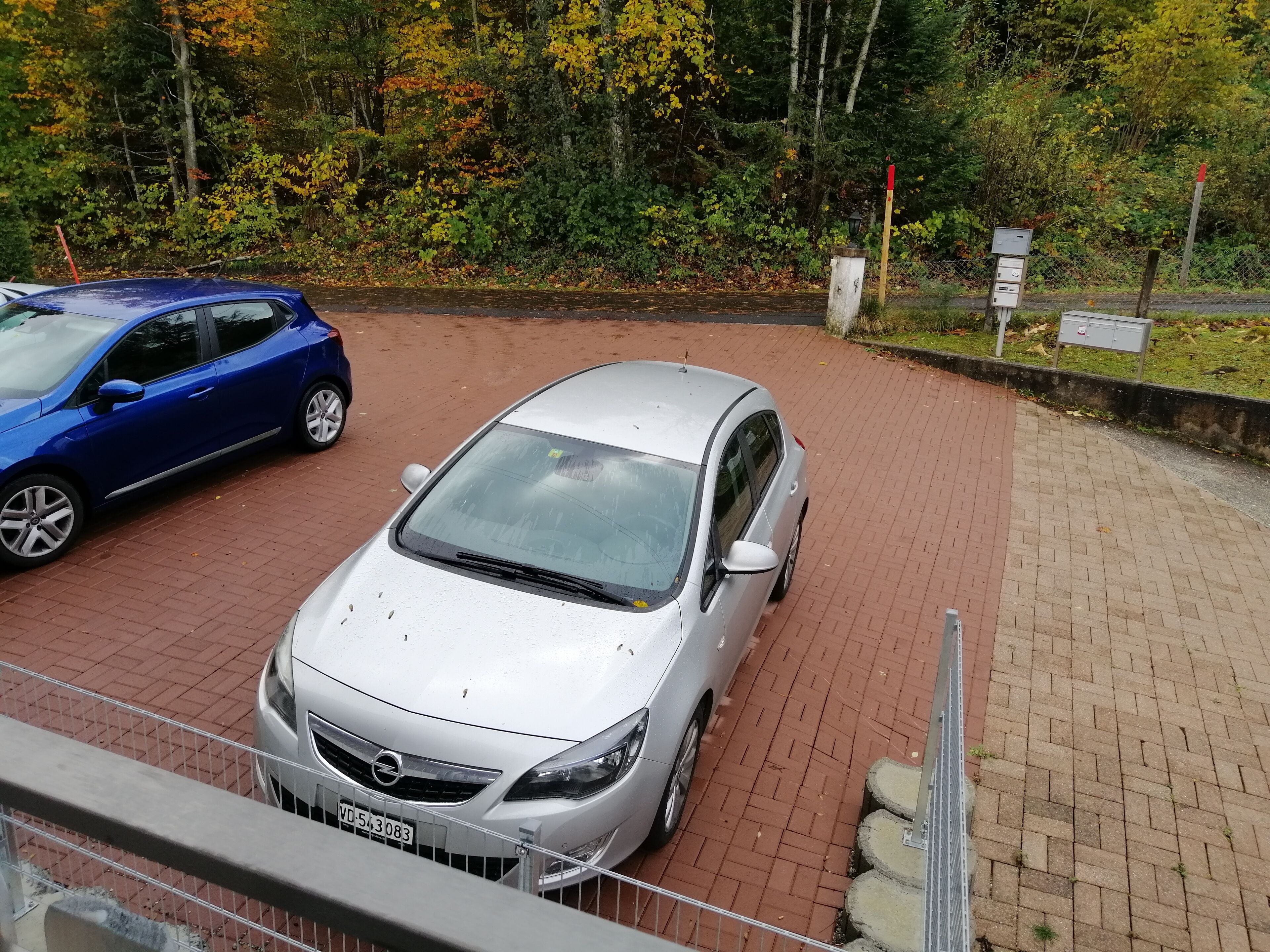 A silver car parked on a paved area with a brick border, surrounded by trees and greenery.
