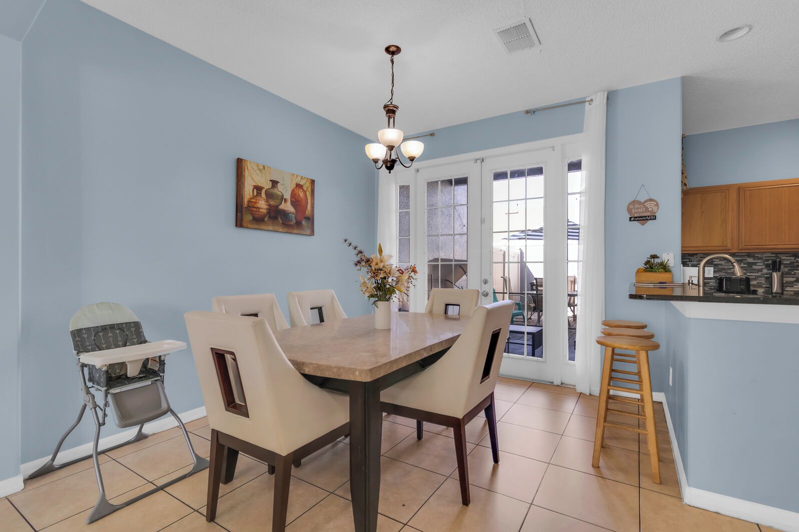 A dining area with a table and chairs, a baby high chair, a chandelier, and a kitchen area with stools.