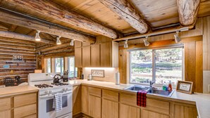 Une cuisine de style rustique, aménagée comme une cabane, avec des meubles en bois, une cuisinière à gaz et une grande fenêtre donnant sur l’extérieur.