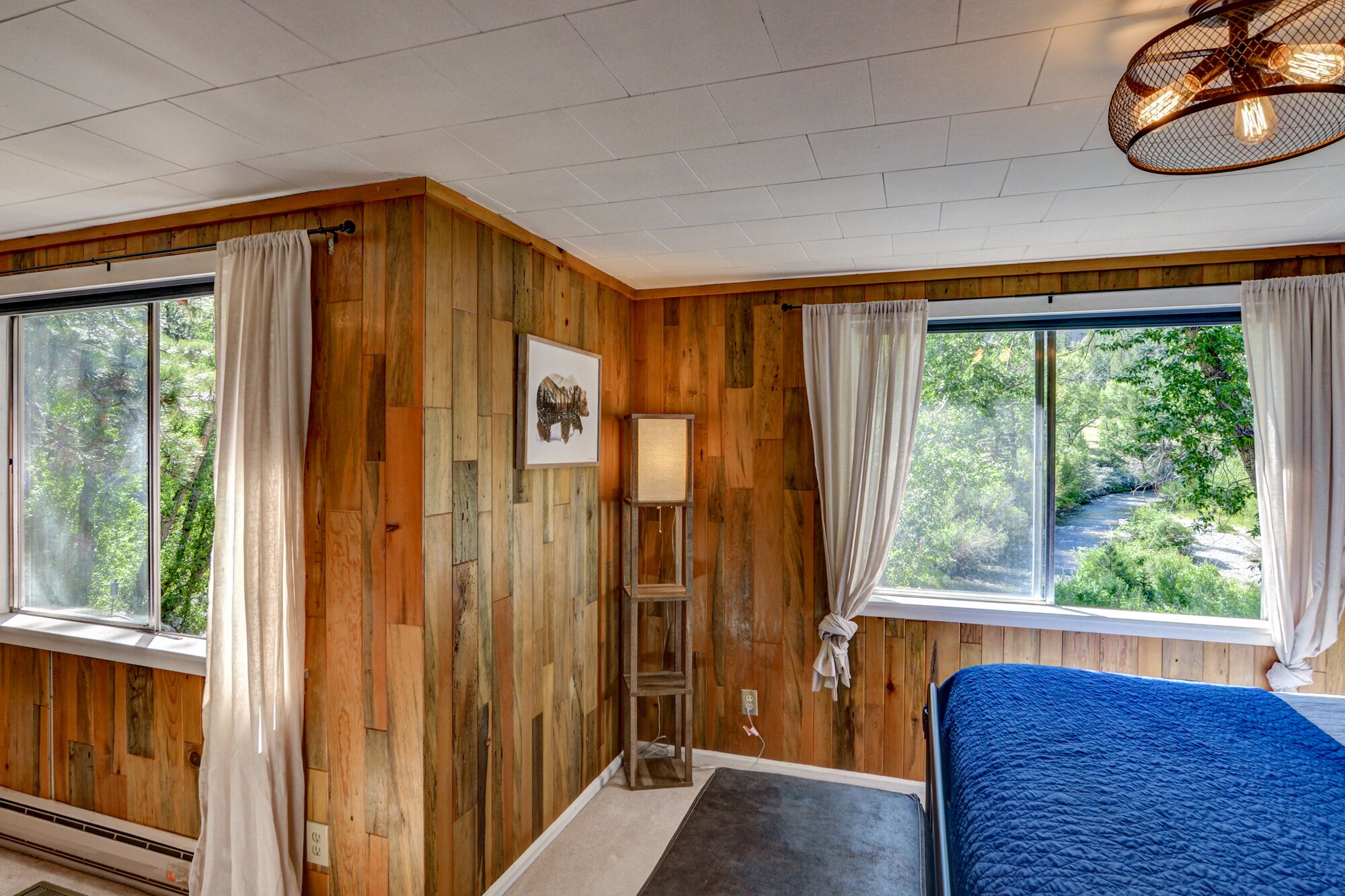Une pièce aux murs recouverts de lambris en bois, un lit avec une couverture bleue, un lampadaire et un tableau accroché au mur.