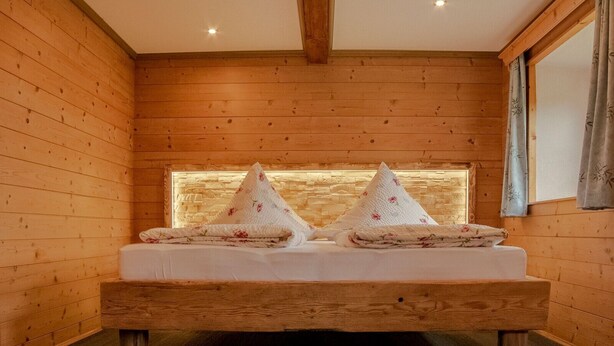 A wooden room with a bed, two pillows, and a window with curtains. - Ferienwohnung , Terr., Küche, 2 Schlaf- u. 1 Wohn-/schlafz., max 4 P (Ruhpolding)