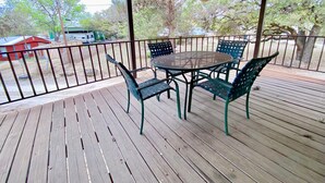 Un espace extérieur abrité, doté d’une terrasse en bois, d’une table et de chaises en métal, et offrant une vue sur un bâtiment rouge et des arbres en arrière-plan.