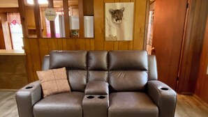 Un canapé en cuir marron avec un seul coussin, une tête d’animal encadrée et une enseigne portant le nom « Wilson », dans une pièce aux murs en bois et dotée d’une fenêtre ornée de rideaux roses.