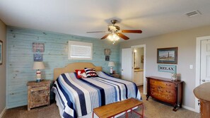 Um quarto com cama, ventilador de teto, mesa de cabeceira de madeira, cômoda e um banco.
