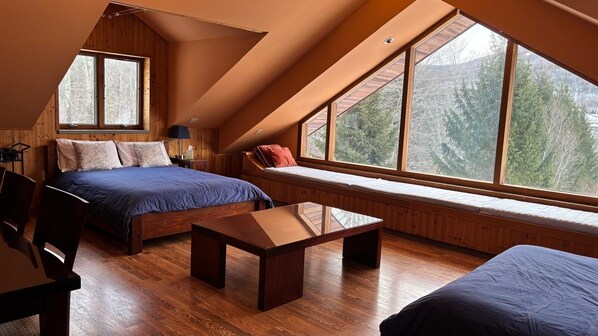 Un dormitorio con techo inclinado, una cama, un banco y un amplio ventanal con vistas a árboles.