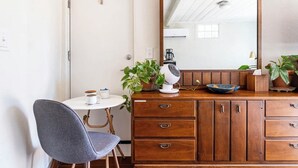 A small, well-lit room with a wooden dresser, a round table with a chair, a mirror, and a potted plant.