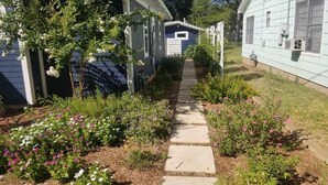 A pathway leading to a small building with a blue door, surrounded by flowering plants and a garden. - In Town. Pet Friendly. Quiet Neighborhood. Queen Bed. Perfect Getaway! (Fredericksburg)