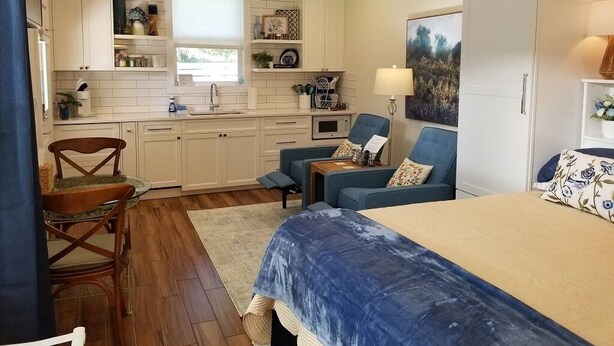 A compact bedroom with a bed, a chair, a small table, and a kitchenette. - In Town. Pet Friendly. Quiet Neighborhood. Queen Bed. Perfect Getaway! (Fredericksburg)