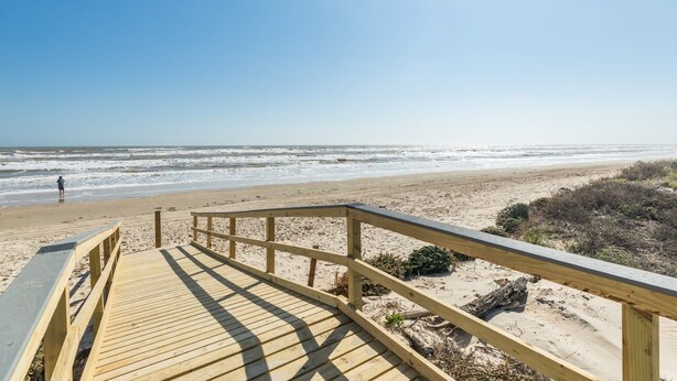 Ein hölzerner Steg führt zu einem Strand bei blauem Himmel.