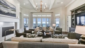 A spacious living room with a fireplace, a large chandelier, and a view of the ocean through large windows. - THE NEW PREMIER BEACHFRONT PLANTATION HOME, LAZY GATOR, IS FINALLY COMPLETE! (St. George Island)
