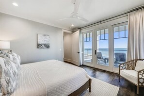 A bedroom with a large bed, a ceiling fan, a view of the ocean through a sliding glass door, and a chair by the door.