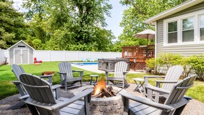 An outdoor patio area with Adirondack chairs, a fire pit, and a pool in the background.