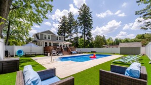 A backyard with a swimming pool, wicker furniture, a wooden deck, and a house in the background.
