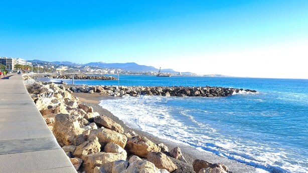 A coastal walkway with a rocky barrier, a clear blue sea, and distant buildings.
