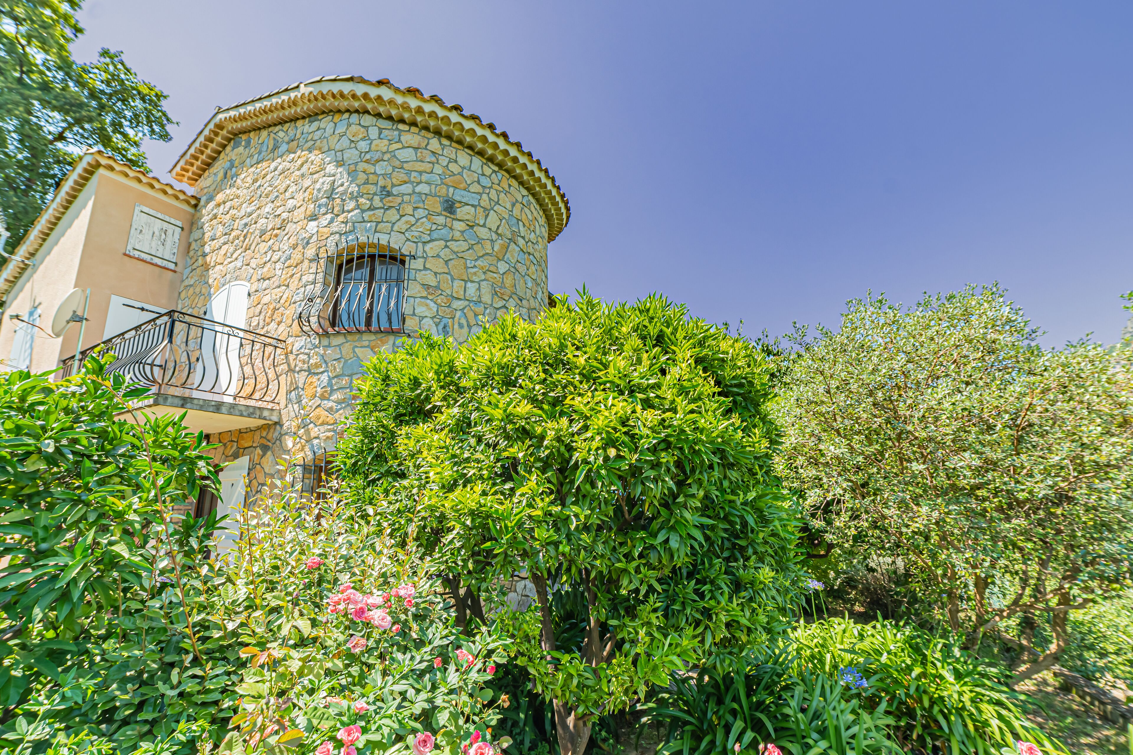 A stone building with a balcony, surrounded by lush greenery and flowering plants.