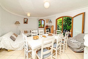 Un espace repas avec une table et des chaises blanches, un canapé et une vue sur la verdure à l’extérieur.