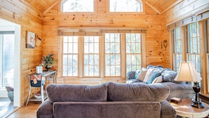 A cozy cabin interior with a wooden ceiling, large windows, a gray sofa, and a lamp on a side table. - On Beautiful White River, Minutes from Sylamore Creek and Blanchard Springs (Mountain View)