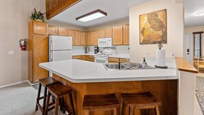 A modern kitchen with wooden cabinets, a white island with stools, a microwave, and a large painting on the wall.
