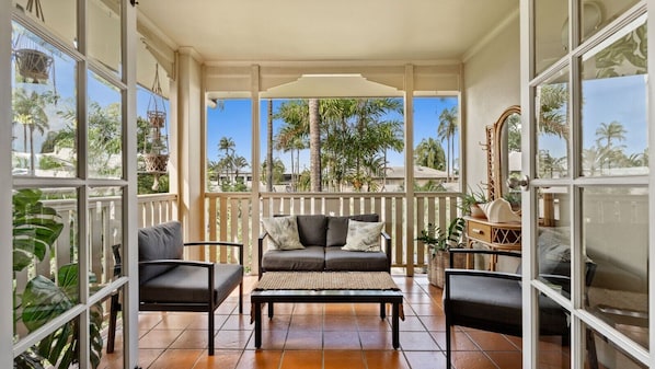 A sunroom with a sofa, chairs, and a coffee table, featuring large windows and a view of palm trees.