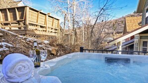 A hot tub with a towel and a bottle, surrounded by a wooden deck and a building.
