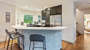A modern kitchen with a central island, dark cabinets, stainless steel appliances, and a view of greenery outside.
