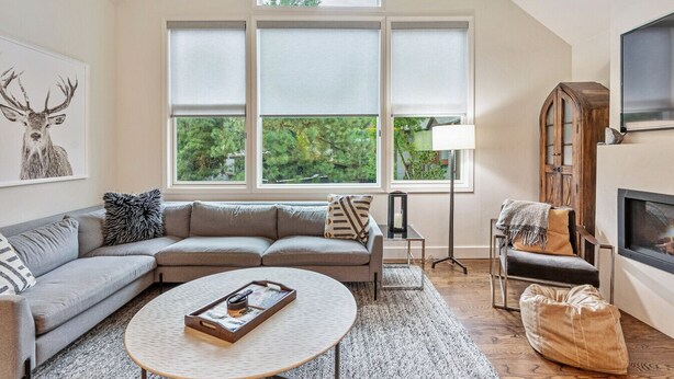 A living room with a sectional sofa, a round coffee table, a fireplace, and large windows with a view of greenery.
