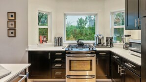 A modern kitchen with a central stove, a microwave, and a refrigerator. There are two windows providing natural light and a view of greenery outside.