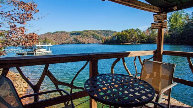 A wooden deck with a table and chairs overlooking a lake, signs indicating White Water Falls and Wrights Creek, and a dock with a boat. - Black Bear Lodge at gorgeous Lake Jocassee (Salem)