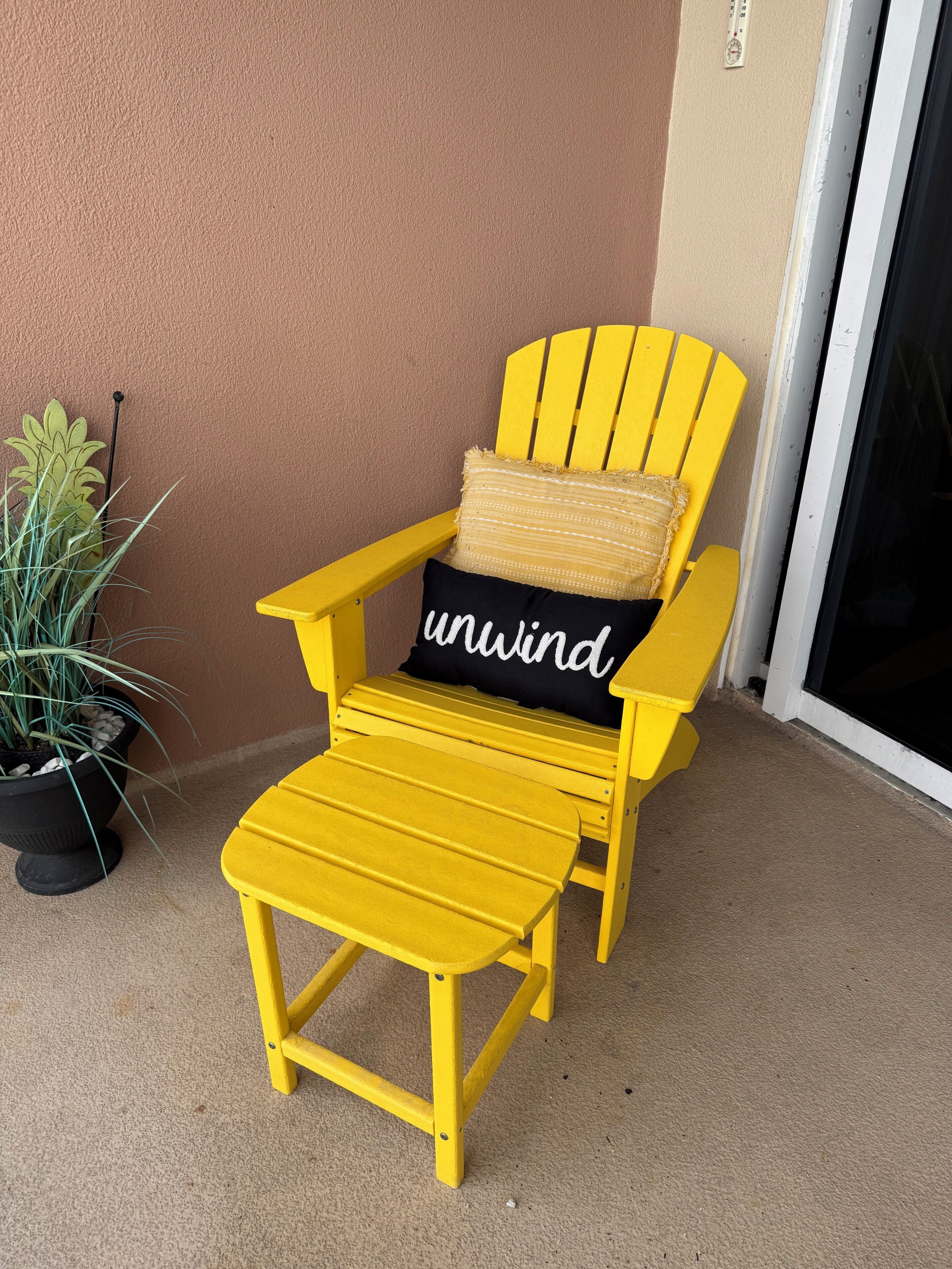 A yellow Adirondack chair with a cushion labeled 'unwind' and a small table beside it, positioned on a balcony with a potted plant nearby.