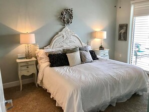 A neatly made bed with white bedding, two bedside tables with lamps, a decorative headboard, and a window with blinds.