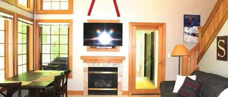 A living room with a fireplace, a television mounted above it, a dining area with a table and chairs, and a staircase leading to another level.
