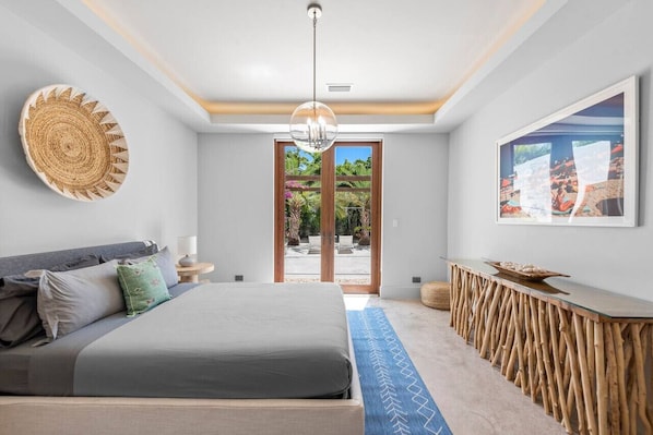 A modern bedroom with a bed, a side table, a woven wall art piece, a glass coffee table, and a large window with a view of greenery.